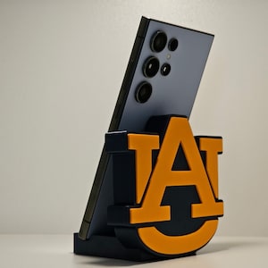 May include: A smartphone is propped up in a black and orange phone stand. The stand features the letters "AU" in orange, with a black outline. The phone is a light blue color and has multiple camera lenses.