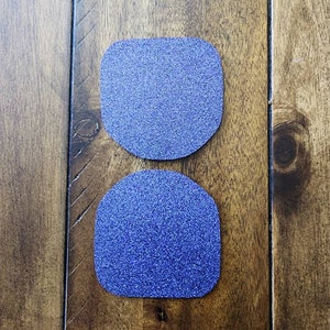 May include: Two blue glittery, rounded-rectangle shapes are displayed on a wooden surface. The shapes are a vibrant blue color and have a textured, sparkling appearance. The wooden surface has a warm, brown tone with visible wood grain.