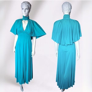 May include: A teal vintage dress with a cape overlay. The dress has a high neckline with a keyhole detail and a flowing, floor-length skirt. The cape sleeves extend outwards, creating a dramatic silhouette. The dress is displayed on a mannequin.