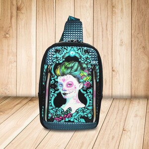 Könnte beinhalten: Schwarze Umhängetasche mit einem Tag der Toten Zuckerschädel-Design. Die Tasche hat ein türkis-schwarzes Farbschema mit einer detaillierten Illustration eines Frauengesichts mit floralen und Schmetterlingsakzenten. Sie hat ein Reißverschluss-Hauptfach und einen gepolsterten Schultergurt.