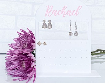 May include: White acrylic earring holder with the name "Rachael" written in pink lettering. The holder has multiple holes for earrings and is displayed on a white surface with a purple flower.