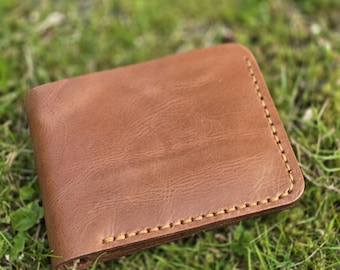 May include: A brown leather wallet with visible stitching around the edges. The wallet is rectangular and appears to be made of high-quality leather. The background is blurred, showing green grass.