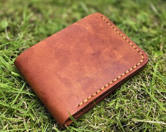 May include: A brown leather wallet with visible stitching around the edges. The wallet is rectangular and appears to be made of a single piece of leather. The wallet is resting on a bed of green grass.