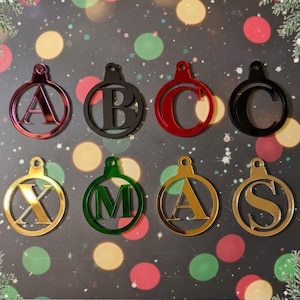 May include: Eight Christmas tree decorations, each shaped like an ornament and featuring a different letter. The decorations are in various colours, including red, black, green, and gold. They have a circular frame and a loop for hanging on the tree.
