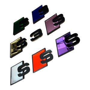 May include: A selection of car emblems in a range of colours: green, blue, pink, gold, red, silver, and purple. Each emblem features a black "S" design, with some featuring a black "3". The emblems have a reflective, glossy finish.