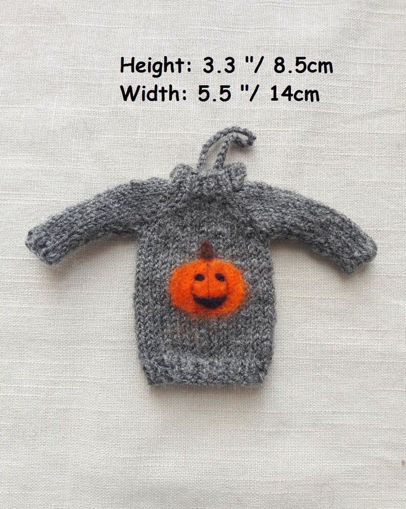 May include: A gray knitted sweater with a felt pumpkin appliqu&eacute;. The pumpkin is orange with a black stem and a smiling face. The sweater measures 3.3 inches tall and 5.5 inches wide.