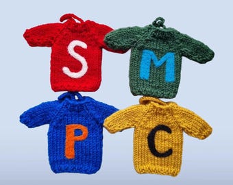 May include: Four miniature knitted sweaters with long sleeves and a small loop at the neck. The red sweater features a white letter S, the green sweater has a blue letter M, the blue sweater displays an orange letter P, and the yellow sweater has a black letter C.