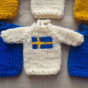 May include: Close-up of several miniature knitted sweaters. The central sweater is white with the Swedish flag, a blue rectangle with a yellow cross. Other sweaters are yellow and blue. The items are likely handmade.
