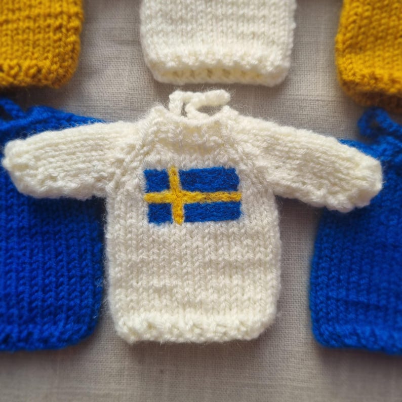 May include: Close-up of several miniature knitted sweaters. The central sweater is white with the Swedish flag, a blue rectangle with a yellow cross. Other sweaters are yellow and blue. The items are likely handmade.