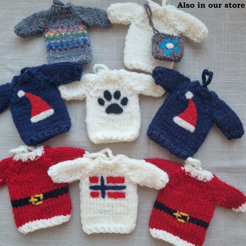 May include: A collection of miniature knitted sweaters in various colors and designs. Some sweaters feature a Santa hat, a paw print, and a Norwegian flag. Others have rainbow stripes or a small purse. The sweaters are likely ornaments or decorative items.