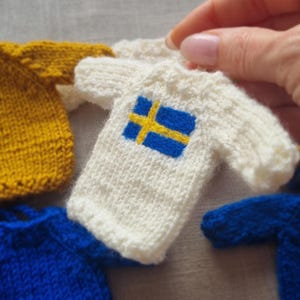 May include: A collection of miniature knitted sweaters in various colors. One white sweater features the blue and yellow cross of the Swedish flag. Other sweaters are yellow and blue. The sweaters are held by a person.