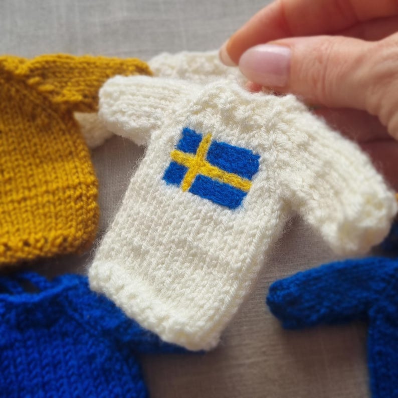 May include: A collection of miniature knitted sweaters in various colors. One white sweater features the blue and yellow cross of the Swedish flag. Other sweaters are yellow and blue. The sweaters are held by a person.
