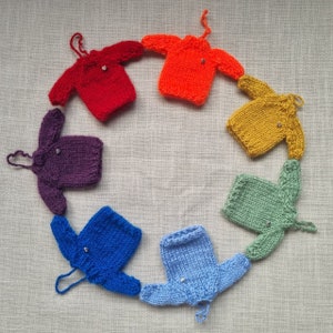 May include: A circular arrangement of seven miniature knitted sweaters in a rainbow of colors: red, orange, yellow, green, light blue, blue, and purple. Each sweater has a small button and a loop for hanging.