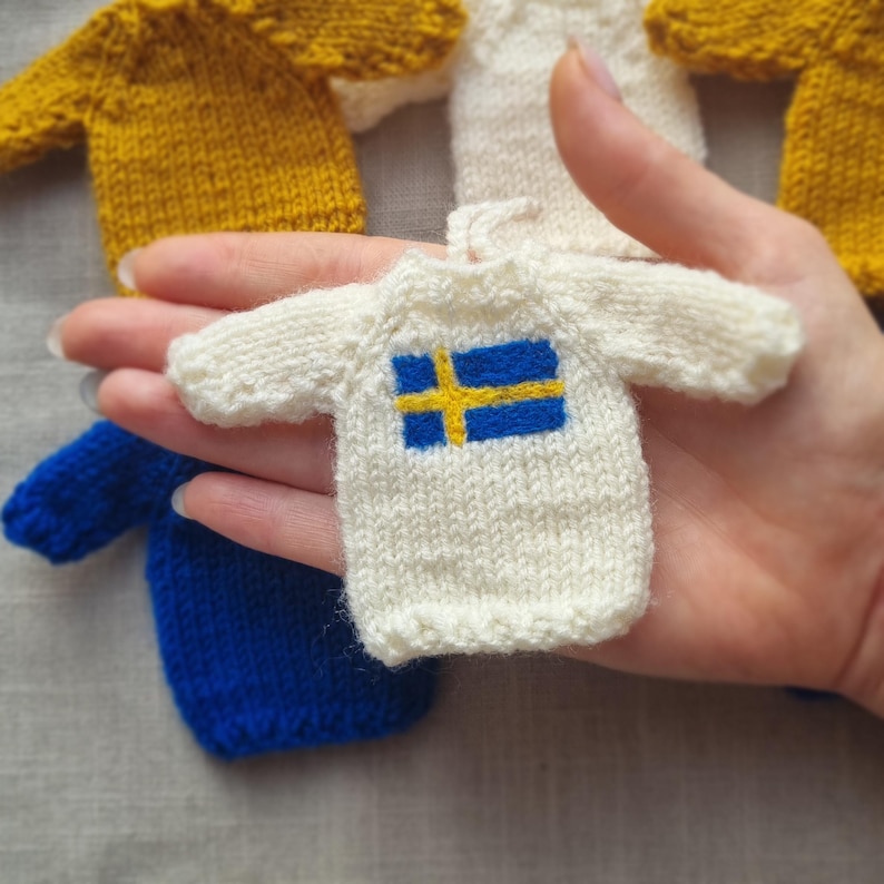 May include: A collection of miniature knitted sweaters in various colors, including white, yellow, and blue. One sweater features the Swedish flag. The sweaters are small, likely for decorative purposes or as doll clothing.