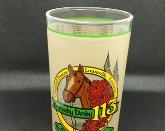 Può includere: Un bicchiere di vetro con un bordo verde e uno sfondo beige. Presenta un'illustrazione colorata di un cavallo, rose e il testo "Churchill Downs, Louisville Kentucky, Kentucky Derby 113, May 2, 1987".