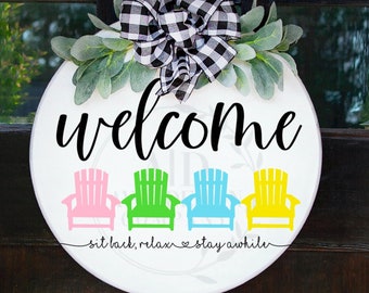 May include: A circular white wooden welcome sign featuring the word "welcome" in black script. Below are four colourful Adirondack chairs in pink, green, blue, and yellow. The sign is decorated with greenery and a black and white buffalo check bow.