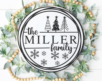 Puede incluir: Un letrero redondo blanco con borde negro y el texto "the MILLER family" con tres árboles de Navidad estilizados y copos de nieve. El letrero está rodeado de una guirnalda de hojas verdes y cuentas beige.