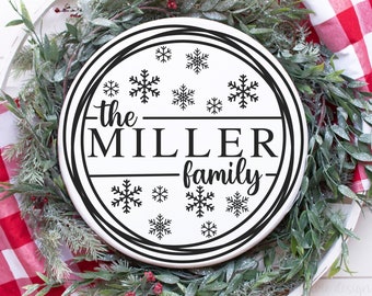 Puede incluir: Un letrero redondo blanco con el texto "the MILLER family" en negro, rodeado de copos de nieve. El letrero está colocado sobre un lecho de vegetación y tela a cuadros rojos y blancos. La imagen también incluye el texto "SVG DXF PNG EPS included".