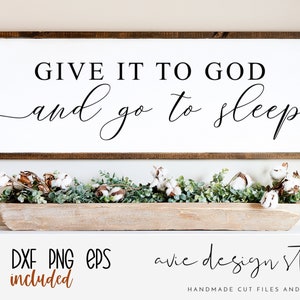 May include: A white sign with a brown wooden frame, featuring the text "GIVE IT TO GOD and go to sleep". The sign is sitting on a white shelf with a wooden bowl filled with greenery and cotton balls.