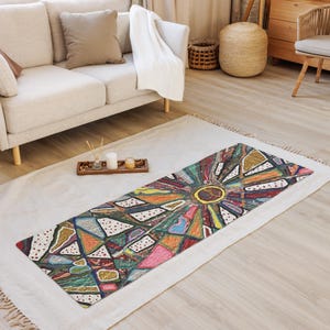 May include: A rectangular yoga mat featuring a vibrant, multi-colored abstract design with a central sun-like motif surrounded by geometric shapes in shades of red, blue, green, yellow, and white, accented with small dark dots throughout the pattern.
