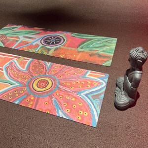 May include: Two rectangular mats featuring a vibrant, hand-painted floral design with large pink, orange, and blue petals, accompanied by a small, grey, seated Buddha figurine. The mats display intricate patterns of dots and lines in various colors.