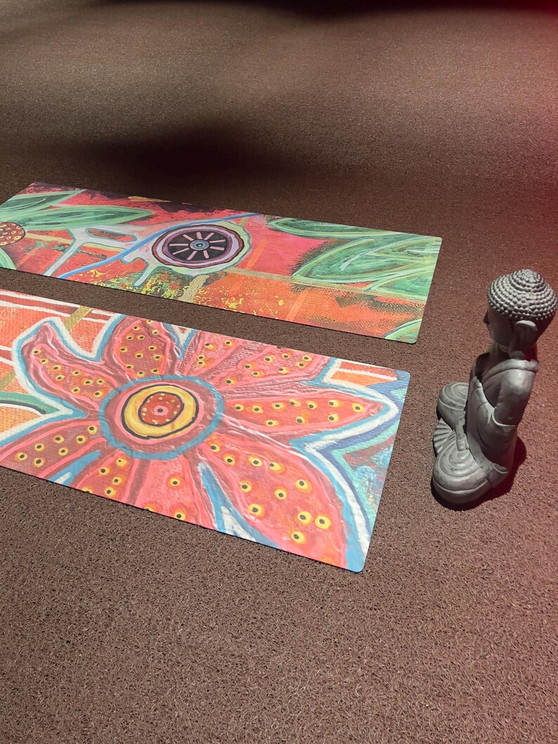 May include: Two rectangular mats featuring a vibrant, hand-painted floral design with large pink, orange, and blue petals, accompanied by a small, grey, seated Buddha figurine. The mats display intricate patterns of dots and lines in various colors.
