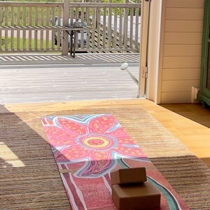 May include: A rectangular yoga mat featuring a large, stylized pink and red flower graphic with yellow accents, laid out on a textured rug. Two brown, rectangular foam yoga blocks are stacked on top of the mat near the bottom edge.