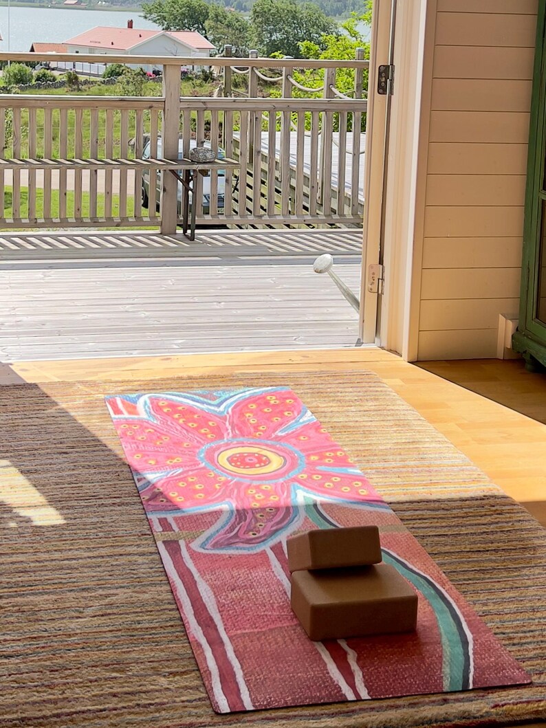 May include: A rectangular yoga mat featuring a large, stylized pink and red flower graphic with yellow accents, laid out on a textured rug. Two brown, rectangular foam yoga blocks are stacked on top of the mat near the bottom edge.