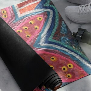 May include: A rectangular yoga mat featuring an abstract design with vibrant pink, blue, and orange brushstrokes. The surface is decorated with multiple yellow circular spots throughout the pink areas. The mat is partially rolled, revealing a solid black underside.