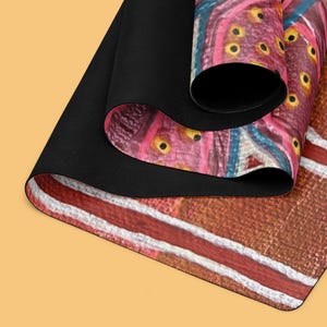 May include: A rectangular mat with a black underside and a rolled top edge. The surface features a vibrant abstract pattern with shades of pink, brown, and blue, accented by small yellow circular details and distinct white horizontal lines.