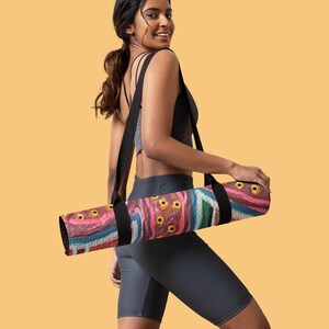 May include: A rolled yoga mat featuring a vibrant abstract pattern with shades of pink, blue, and brown, accented by yellow circular motifs. The mat is equipped with a black carrying strap that loops over the shoulder for transport and storage.