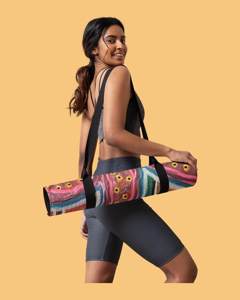 May include: A rolled yoga mat featuring a vibrant abstract pattern with shades of pink, blue, and brown, accented by yellow circular motifs. The mat is equipped with a black carrying strap that loops over the shoulder for transport and storage.
