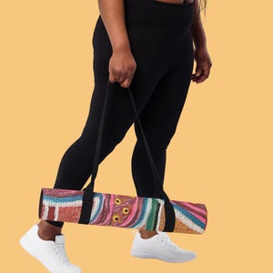 May include: A rolled yoga mat featuring a vibrant, abstract pattern with swirling lines and circular shapes in shades of pink, blue, brown, and white. The mat is equipped with a black carrying strap attached to the center for easy transport.