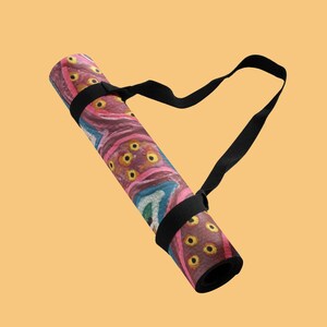 May include: A rolled yoga mat featuring a vibrant, abstract pattern with shades of pink, purple, and teal, accented by yellow circular motifs. The mat is secured with a black adjustable carrying strap that loops around the exterior for transport.