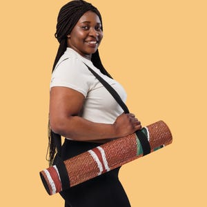 May include: A rolled yoga mat featuring a textured brown surface with white and red horizontal stripes and green accents. The mat is secured with a black carrying strap that loops over the shoulder, allowing for easy transport of the exercise equipment.