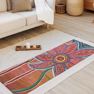 May include: A rectangular area rug featuring a large, stylized flower design with pink, red, and yellow petals adorned with small dots. The background of the rug displays horizontal bands of orange, brown, and white, with a green stem extending downward.