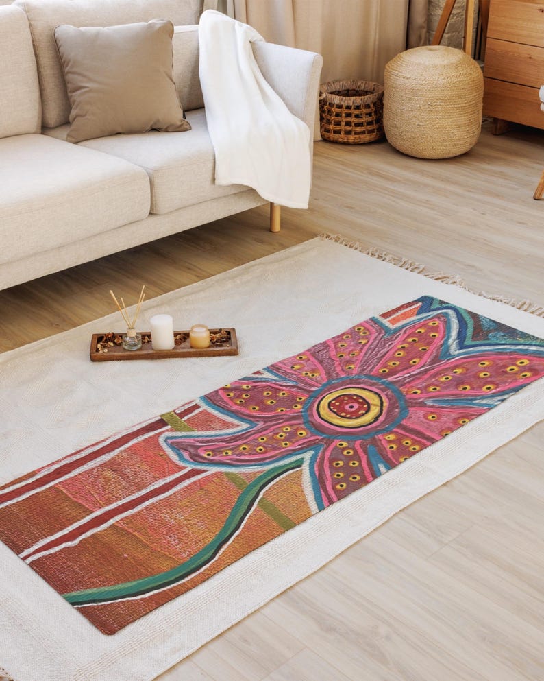 May include: A rectangular area rug featuring a large, stylized flower design with pink, red, and yellow petals adorned with small dots. The background of the rug displays horizontal bands of orange, brown, and white, with a green stem extending downward.