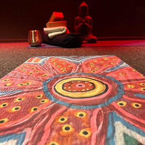 May include: A yoga mat featuring a vibrant, hand-painted floral pattern in shades of red, pink, yellow, and blue with circular motifs. In the background, a dark room contains a yoga bolster, two orange blocks, a white towel, and a Buddha statue.