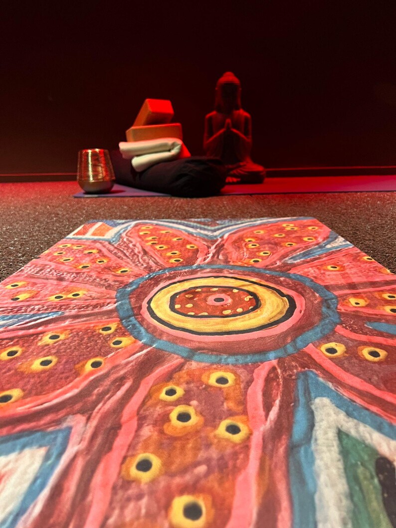 May include: A yoga mat featuring a vibrant, hand-painted floral pattern in shades of red, pink, yellow, and blue with circular motifs. In the background, a dark room contains a yoga bolster, two orange blocks, a white towel, and a Buddha statue.