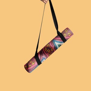 May include: A rolled yoga mat featuring a vibrant, abstract pattern with shades of deep red, pink, blue, and yellow circular motifs. The mat is suspended by a black adjustable carrying strap, showcasing a textured surface and a cylindrical shape.