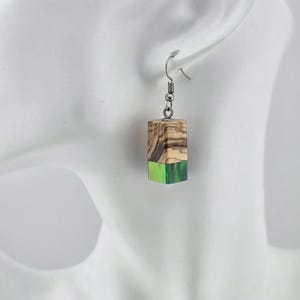 May include: A pair of rectangular earrings with a wooden top and a bright green bottom. The wood has a natural grain pattern with shades of brown and tan. The earrings are suspended from silver-colored fish hook ear wires.