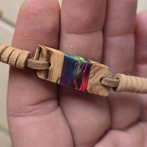 May include: A brown leather bracelet with a wooden and resin inlay. The inlay has a colorful iridescent finish with shades of blue, green, and red.