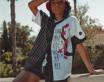 Puede incluir: Una persona lleva una camiseta de béisbol negra y blanca con un diseño japonés y flores rosas. La camiseta tiene un patrón de rayas blancas y negras en un lado y un lado blanco con un círculo negro y texto japonés que dice "Bebe tus sueños". La persona lleva un sombrero de cubo rojo y negro y pantalones cortos negros.
