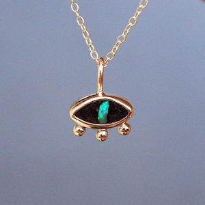 Pode incluir: Um colar de corrente dourada com um pingente em forma de olho. O pingente tem um centro escuro de formato oval com uma incrustação vertical verde iridescente, emoldurado por uma borda dourada com três pequenas esferas douradas na base.