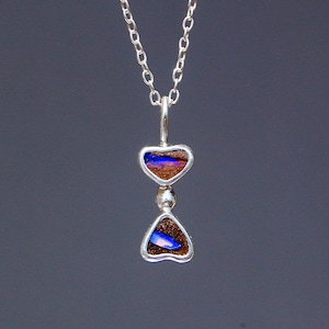 May include: A silver pendant necklace featuring two stacked, heart-shaped settings. Each setting contains a dark brown stone inlay with a vibrant, iridescent blue flash. The pendant hangs from a delicate silver chain with small, uniform links.