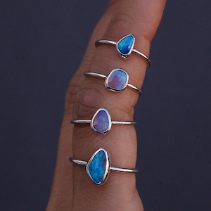 Könnte beinhalten: Vier silberne Ringe mit ovalen, schillernden Opalsteinen in Blau- und Lilatönen. Die Ringe werden auf einem Finger vor dunklem Hintergrund präsentiert. Die Opalsteine haben eine einzigartige, unregelmäßige Form.