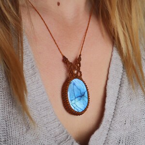 May include: A brown cord necklace with a blue and white oval labradorite gemstone pendant. The pendant is surrounded by a brown macrame design.
