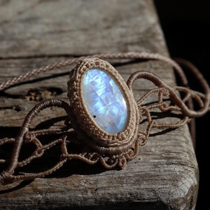 May include: A handcrafted necklace featuring a large, oval moonstone pendant. The moonstone displays a shimmering, iridescent blue and white color. The pendant is set in a woven, beige macrame design, with a matching cord.