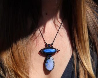 Könnte beinhalten: Eine handgefertigte Halskette mit einem blau-weißen ovalen Anhänger, einem blauen ovalen Stein und einem blauen Stein in der Mitte. Die Halskette hängt an einer schwarzen Kordel. Der Schmuck wird auf einem dunkelblauen Oberteil getragen.