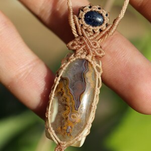 K&ouml;nnte beinhalten: Handgefertigte Makramee-Halskette mit einem polierten, oval geformten Stein mit braunen, gelben und wei&szlig;en Mustern. Der Stein ist in einem beigefarbenen Makramee-Gewebe gefasst, mit einem kleinen blauen Stein-Akzent oben.